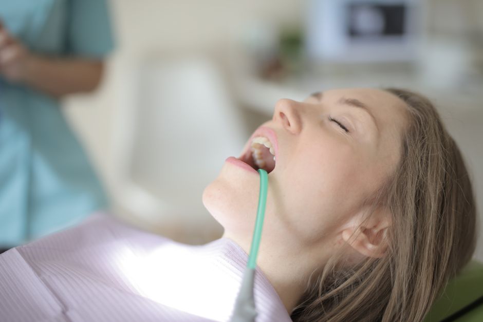 Side view of young female patient with saliva ejector in opened mouth sitting in dental chair while preparing for treatment in modern dental clinic