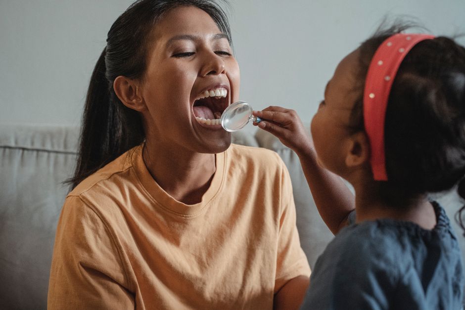 Crop ethnic girl checking mothers teeth with magnifying glass while playing dentist game together at home