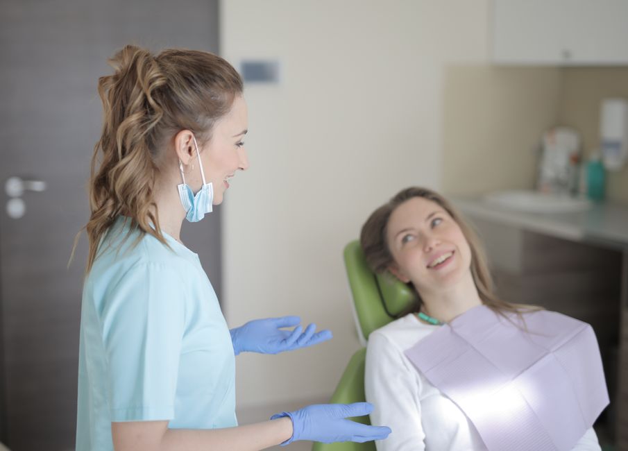Positive young female dentist in latex gloves and uniform talking with young patient while sitting near chair and preparing for treatment in modern dental room in hospital