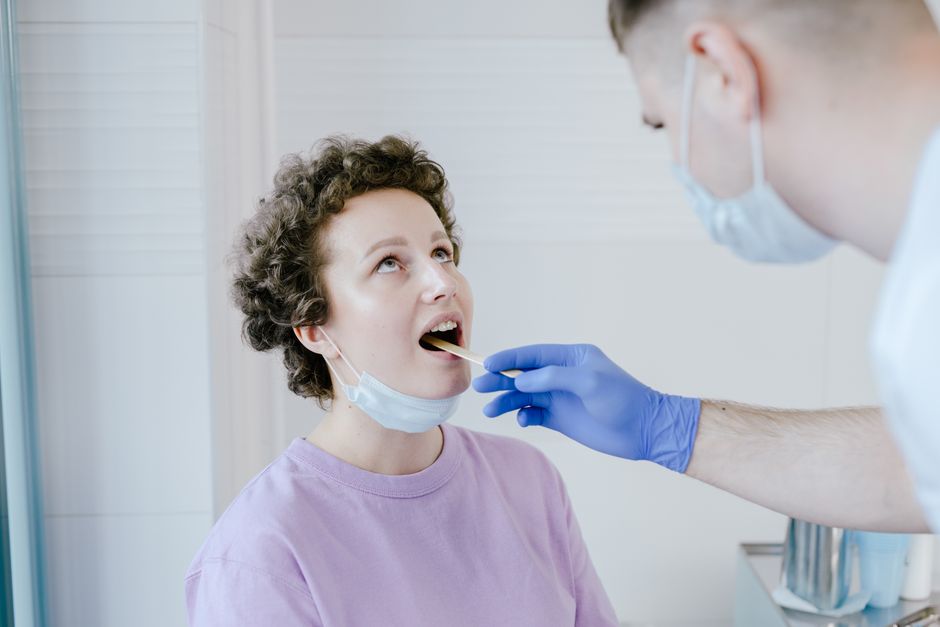 A doctor performs an oral exam on a patient wearing gloves and mask in a clinic.