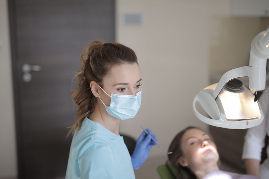 Young female dentist in medical mask and latex gloves wearing uniform while working with patient in modern dental room in hospital