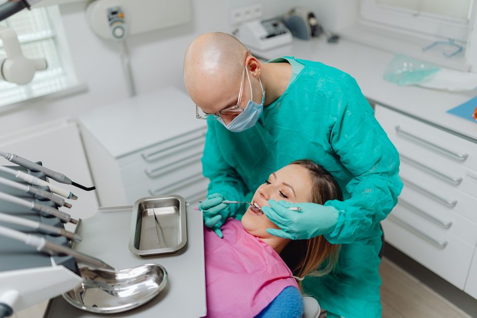 Bald dentist in green scrubs examining a woman's teeth in a modern clinic.