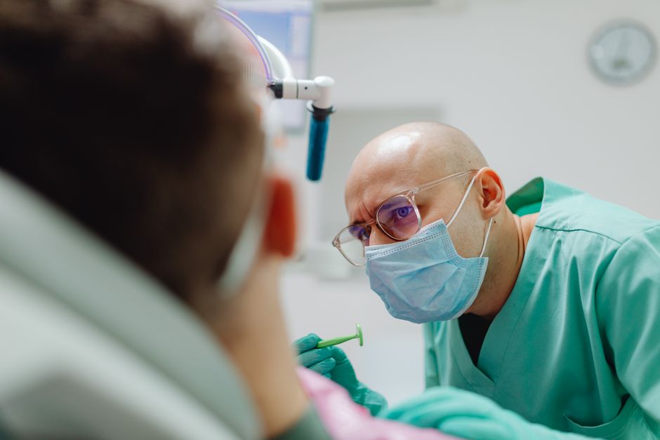 A dentist in a surgical mask examines a patient in a clinical setting.