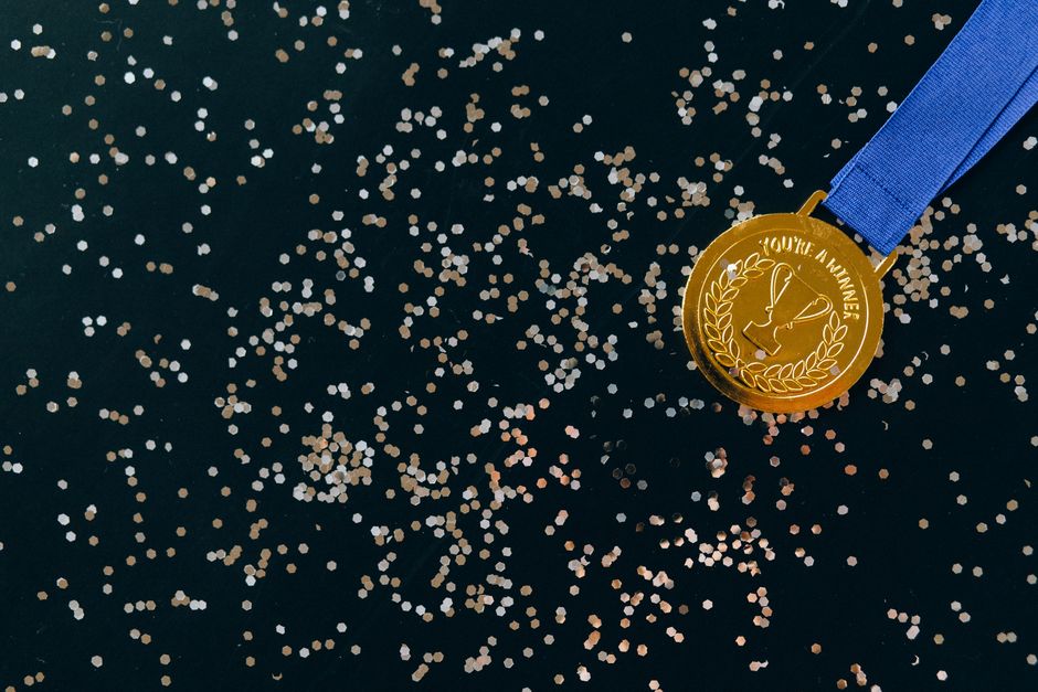 Close-up of a gold medal with blue ribbon and confetti on a black surface, symbolizing victory.