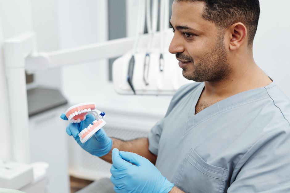 Dentist wearing scrubs and gloves demonstrates dental care with a teeth model.