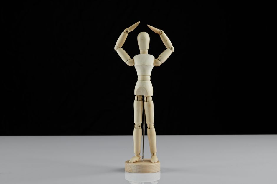 A wooden mannequin with arms raised displayed on a reflective surface.