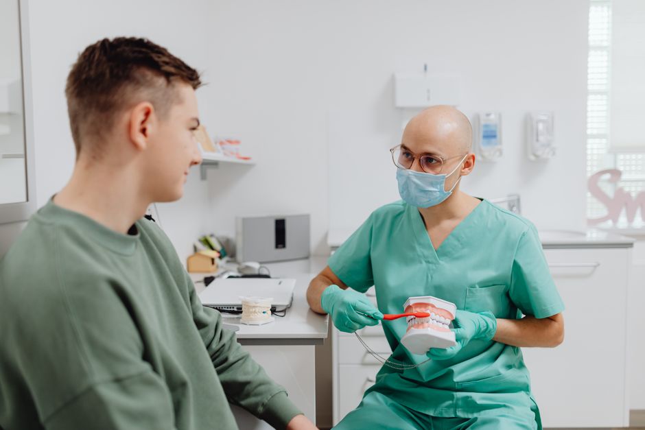 A dentist in scrubs teaches a young man oral hygiene using a dental model in a bright clinic.
